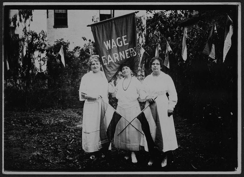 Three women stand holding a sign that says "wage earners" as well as two women's suffrage flags with womens suffrage flags behind them