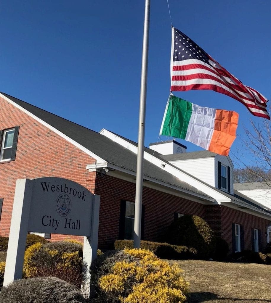 The American flag flies over the Irish flag in front of Westbrook City Hall. A Sign that says "Westbrook city hall" is on the left. 
